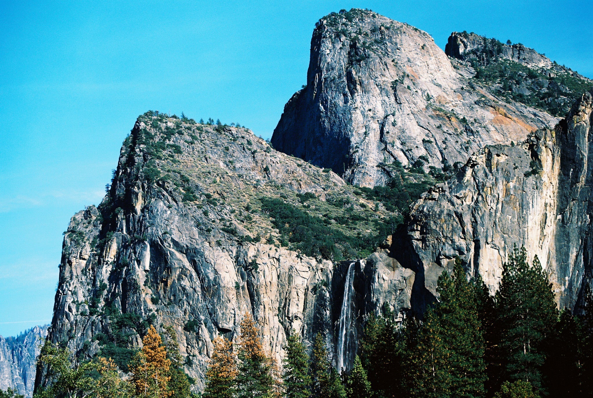 A rocky mountain with a clear sky background and some trees in the foreground.